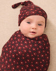 A baby swaddled and wearing a matching knit hat in a dark pink floral pattern, lying on a beige textured blanket.