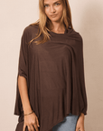 A woman wearing a draped, oversized brown top with three-quarter sleeves and an asymmetrical hem, paired with light blue jeans, against a plain beige background.