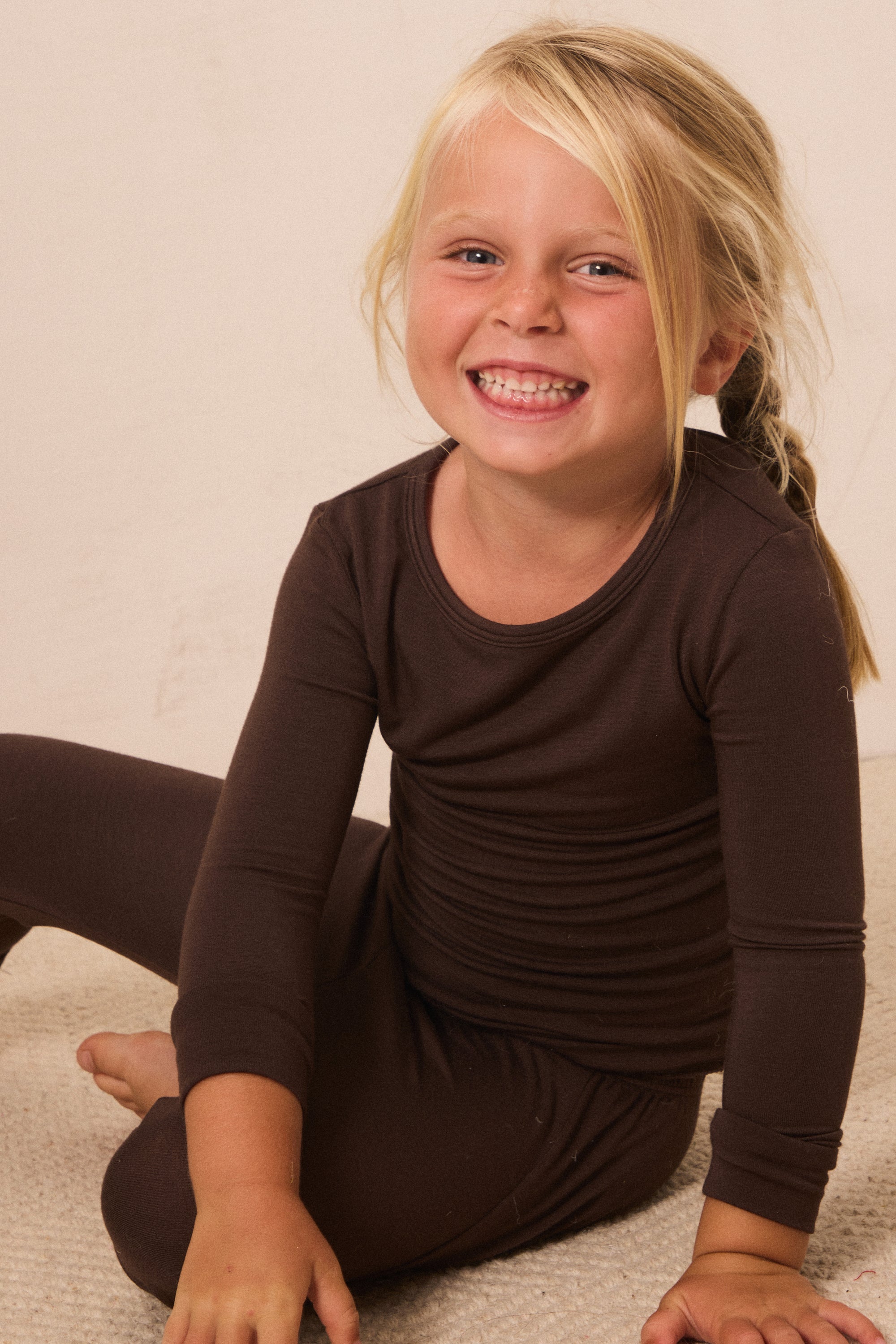 A young blonde girl smiling while wearing a dark brown long-sleeve top and matching dark brown pants, sitting on a light beige carpet against a plain beige wall.