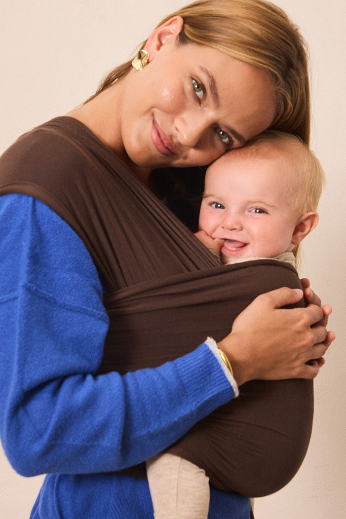A woman wearing a bright blue sweater carries a smiling baby in a dark brown wrap carrier against a plain beige background.