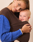 A woman wearing a bright blue sweater carries a smiling baby in a dark brown wrap carrier against a plain beige background.