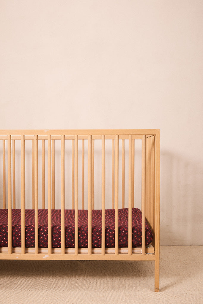 A light natural wood crib with vertical slats, shown empty with no mattress or bedding, set against a plain beige wall and textured beige floor.