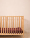 A light natural wood crib with vertical slats, shown empty with no mattress or bedding, set against a plain beige wall and textured beige floor.