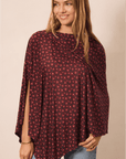 A woman wearing a dark pink flower patterned poncho top with a small white and red floral pattern, paired with light blue jeans, against a solid beige background.