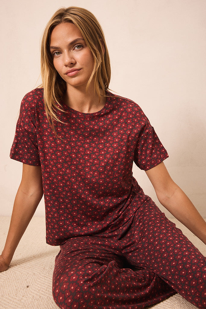 A woman wearing a matching short-sleeve t-shirt and wide-leg pants pajama set in a deep red color with a small repeating yellow floral pattern, sitting against a plain beige background.