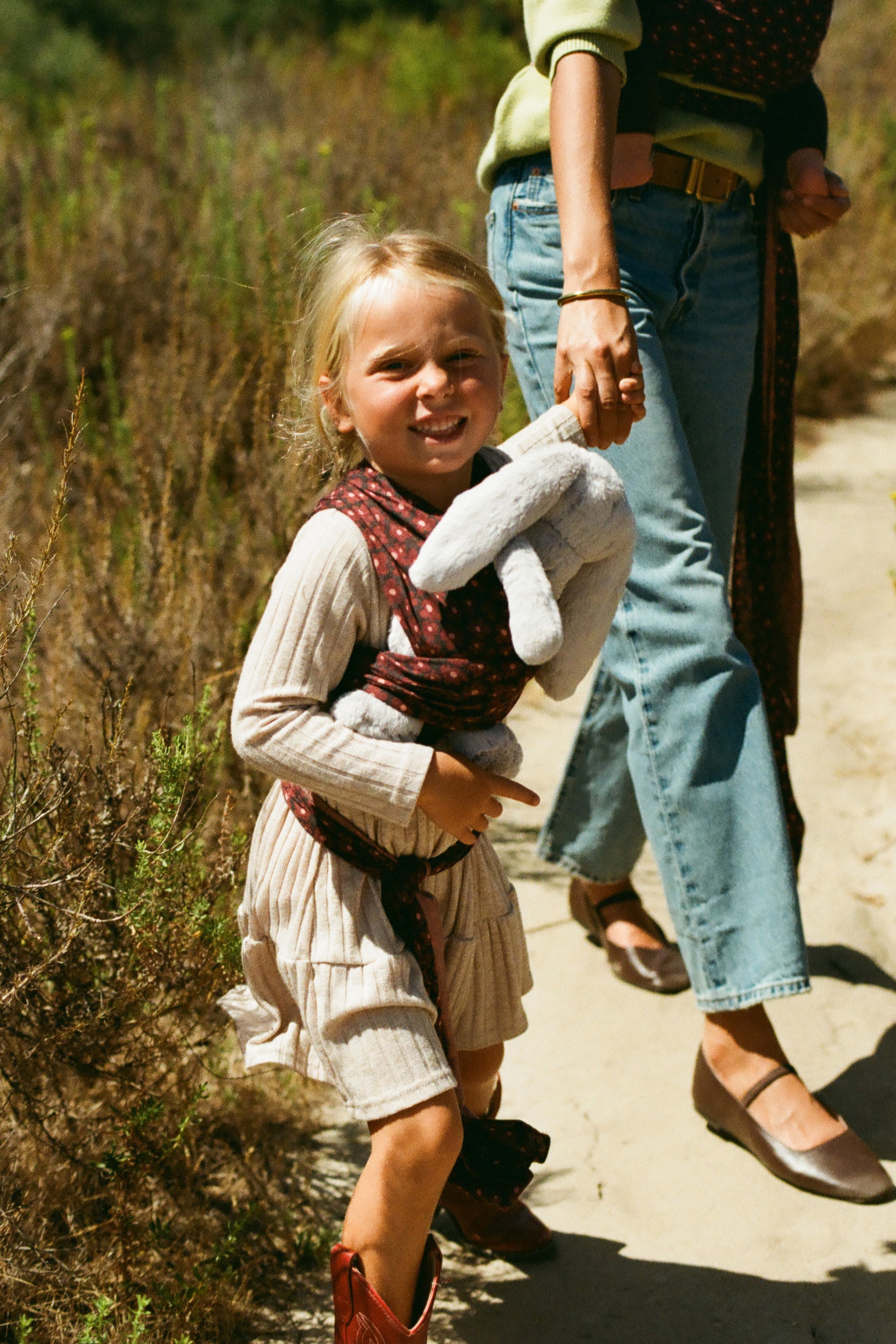 A young girl in a cream ribbed dress and patterned vest carries a gray stuffed bunny while walking on a dirt path with a woman in jeans and a green top beside her.