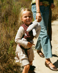 A young girl in a cream ribbed dress and patterned vest carries a gray stuffed bunny while walking on a dirt path with a woman in jeans and a green top beside her.
