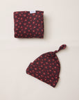 A folded matching baby set consisting of a soft cloth wrap and a knotted beanie hat, both featuring a dark red and navy blue floral pattern, laid out on a light beige background.