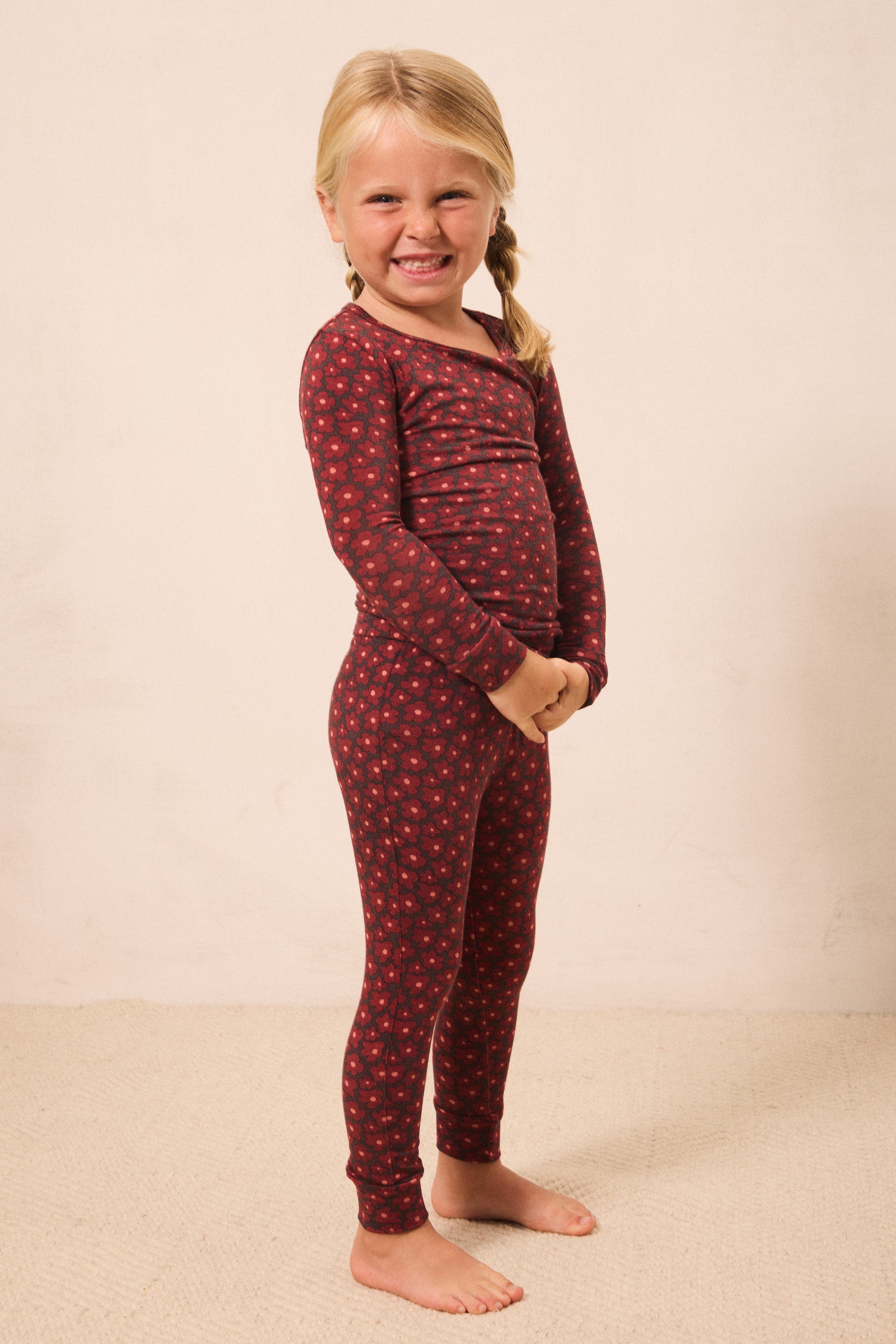 A young girl with blonde braids stands barefoot against a plain beige wall, wearing a long-sleeve one-piece pajama set in dark red with a small repeating floral pattern.