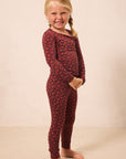 A young girl with blonde braids stands barefoot against a plain beige wall, wearing a long-sleeve one-piece pajama set in dark red with a small repeating floral pattern.