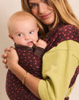 A woman wearing a green and burgundy polka-dot baby wrap carrier holds a baby against her chest. The baby is also wearing a matching brown wrap.