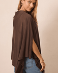 A woman wearing a brown draped poncho-style top with side slits and blue jeans, photographed against a plain beige studio background.