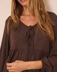 A woman wearing a dark brown long-sleeve blouse with a tie-neck detail and a gathered waist, shown in a close-up shot against a plain beige background.
