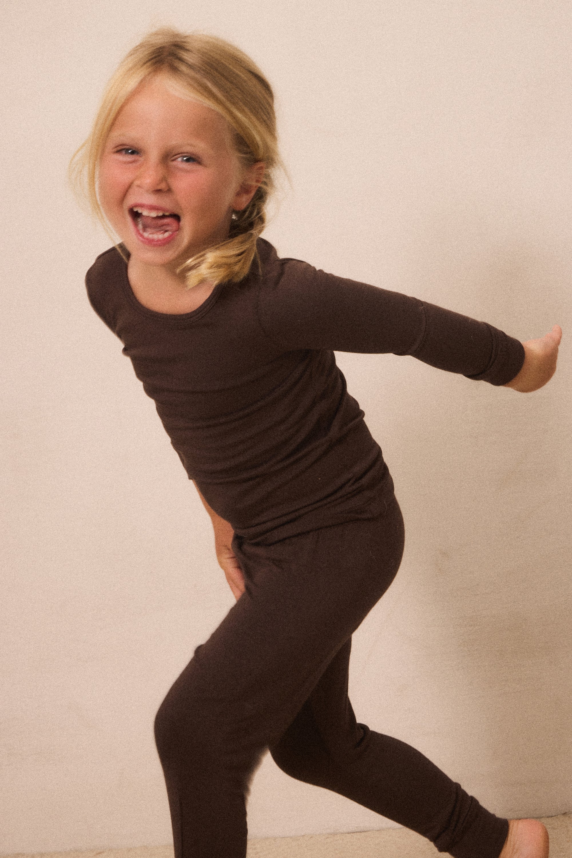 A young blonde girl wearing a dark brown long-sleeve top and matching dark brown leggings, captured in a playful pose against a plain beige wall.