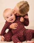 Two babies wearing matching dark red and black patterned long-sleeve footless onesies, with one baby hugging and kissing the other on the cheek, on a beige carpeted floor.