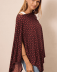 A woman wearing a dark pink and flower patterned poncho top with blue jeans, photographed against a plain beige studio background.