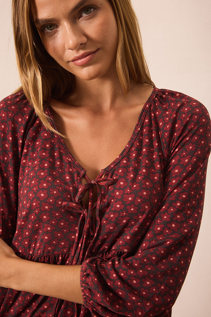 A close-up of a woman wearing a dark pink blouse with a small red and pink floral pattern and a tie detail at the V-neck.