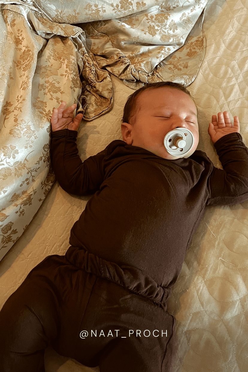 A sleeping newborn baby wearing a dark brown long-sleeve romper and tights, lying on a beige quilted bed with a patterned gold and beige blanket. The baby has a white pacifier in their mouth, and the handle of the photo is signed @NAAT_PROCH.