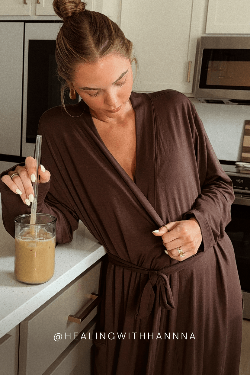 A woman in a brown wrap robe leans on a kitchen counter with a glass of iced coffee, wearing gold hoop earrings and a ring. The text '@HEALINGWITHHANNA' is visible at the bottom.