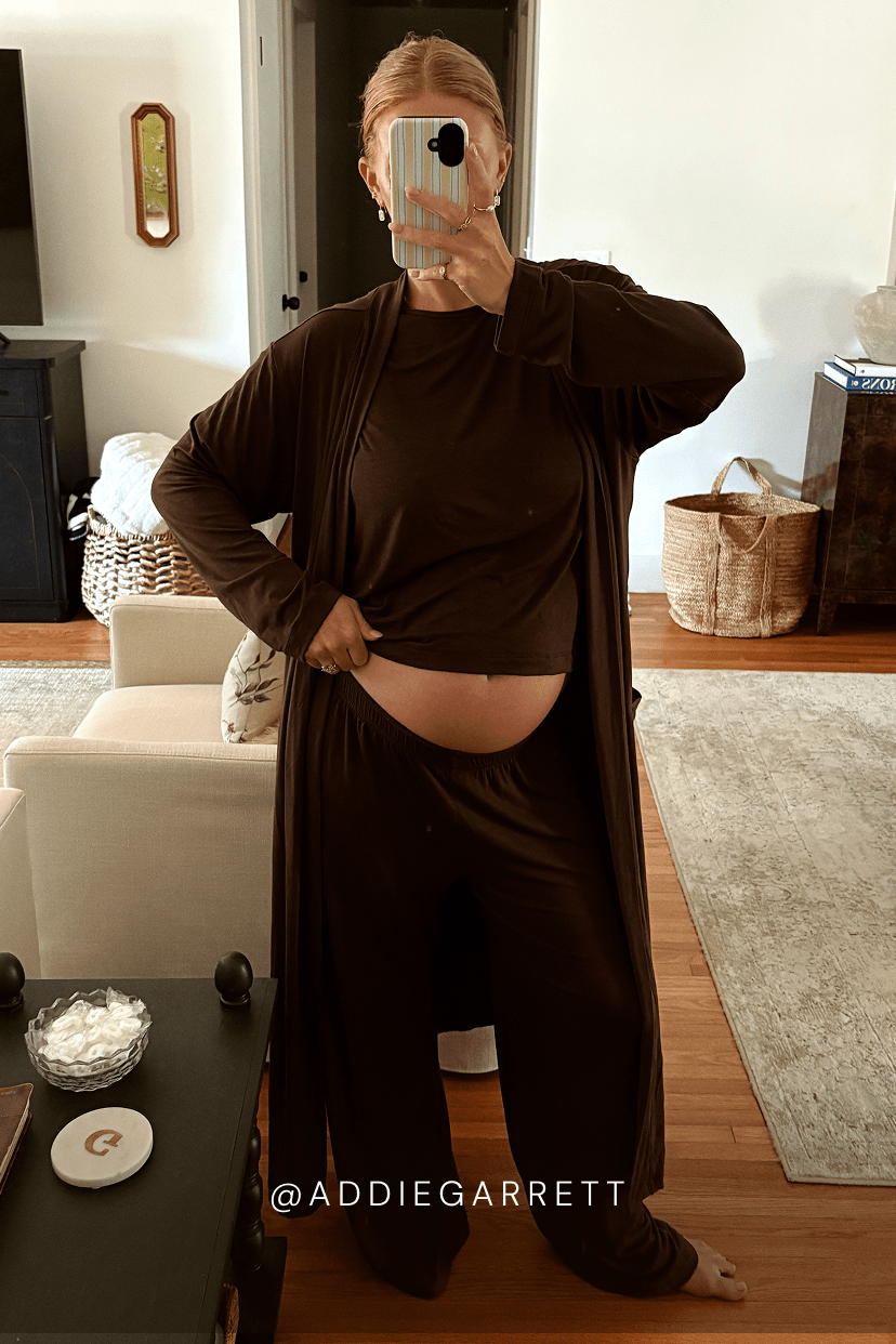A pregnant woman takes a mirror selfie in a living room, wearing a dark brown long-sleeve crop top, matching wide-leg pants, and a long open-front cardigan. She is holding a phone to take the photo, with a visible belly.