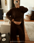 A pregnant woman takes a mirror selfie in a living room, wearing a dark brown long-sleeve crop top, matching wide-leg pants, and a long open-front cardigan. She is holding a phone to take the photo, with a visible belly.