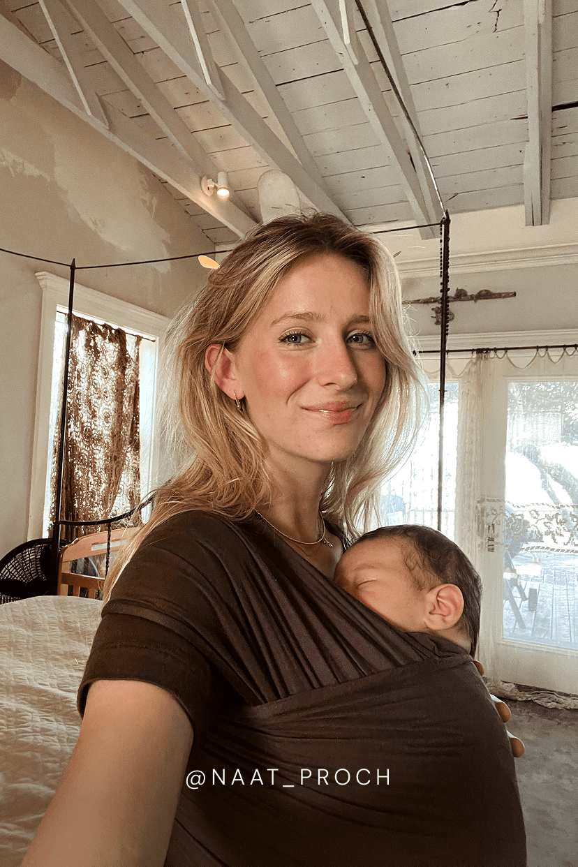 A woman wearing a dark brown baby wrap carrier with a sleeping newborn baby, photographed in a home interior with high white ceiling beams and windows in the background.