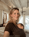 A woman wearing a dark brown baby wrap carrier with a sleeping newborn baby, photographed in a home interior with high white ceiling beams and windows in the background.