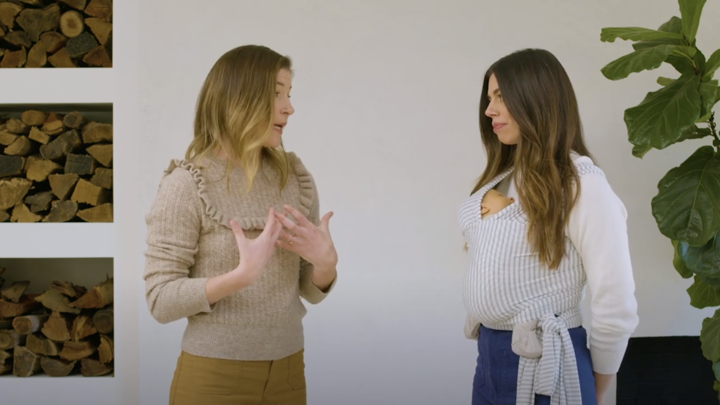 Beige ruffled knit sweater on woman talking to woman wearing white top with gray striped baby wrap carrier and doll.