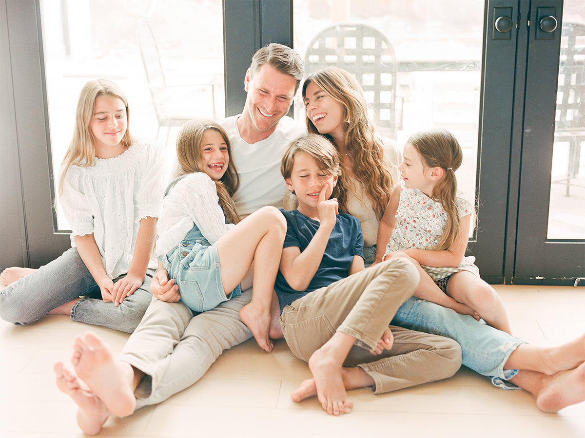 Casual family sitting on wooden floor by glass doors, wearing light-colored casual clothes, smiling and laughing together