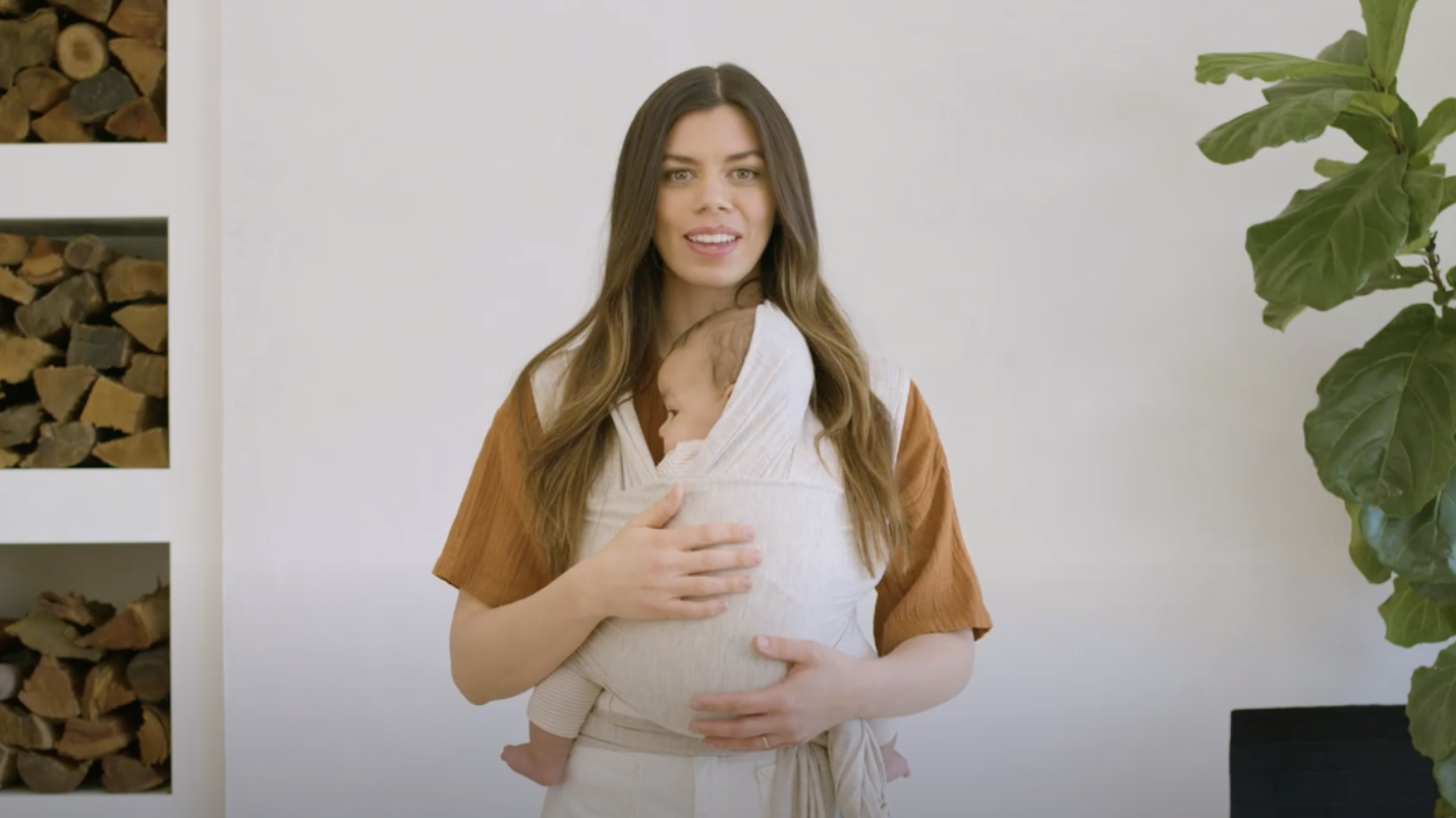 Woman wearing white baby wrap carrier holding a baby with bundled head against a white wall background
