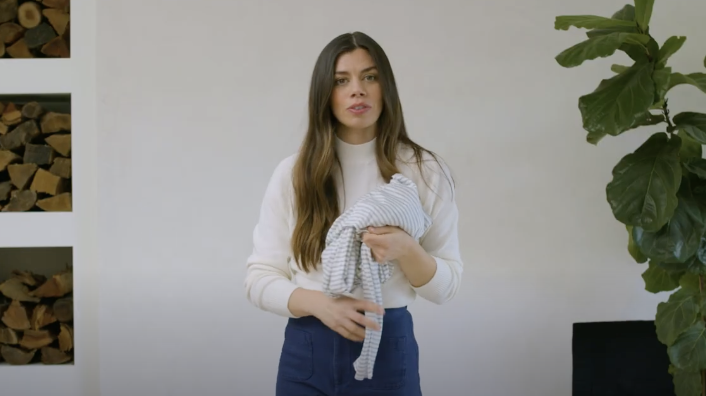 Woman wearing white sweater and blue pants holding a gray and white striped fabric or garment indoors near plant and firewood.