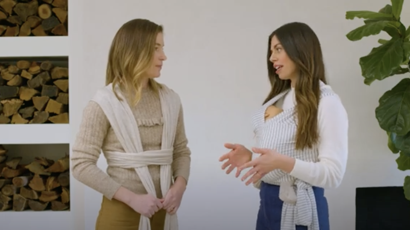 Two women demonstrating baby wrap carriers, one beige and one white with gray stripes, indoors near woodpile and plant.