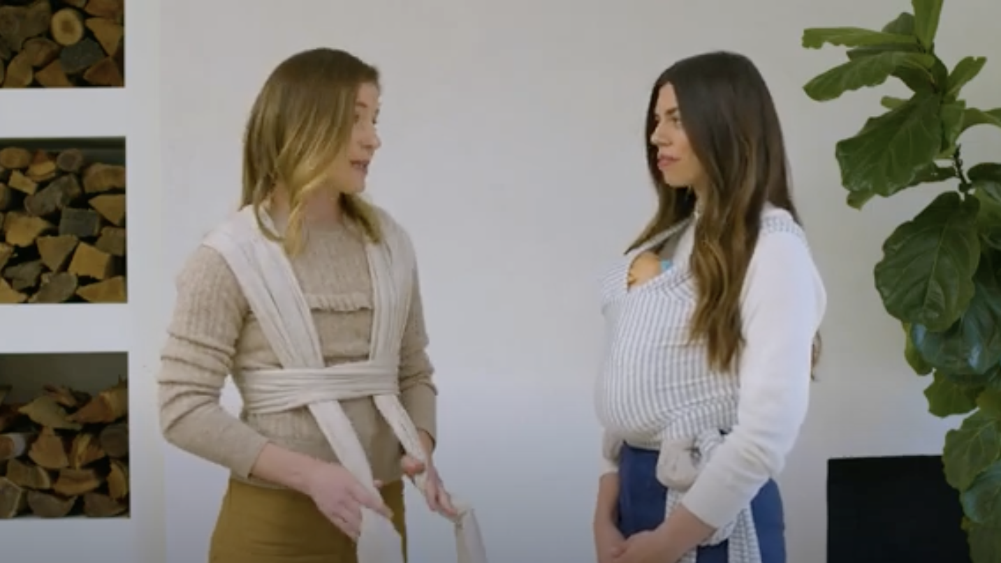 Two women demonstrating beige and white striped baby carriers with babies inside, standing indoors near a plant.