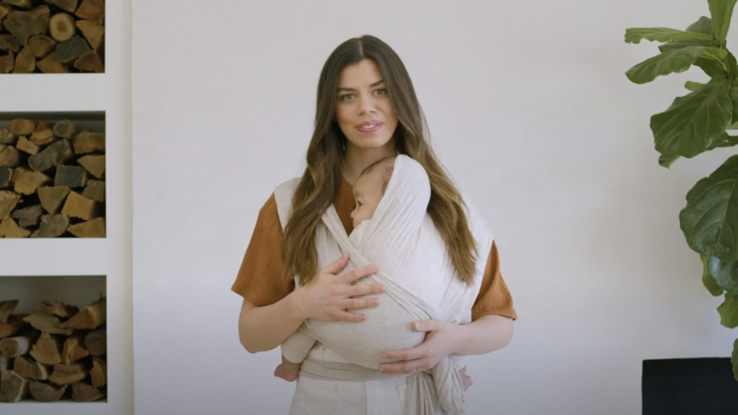 Woman wearing a light beige baby wrap carrier holding a baby dressed in matching beige outfit indoors