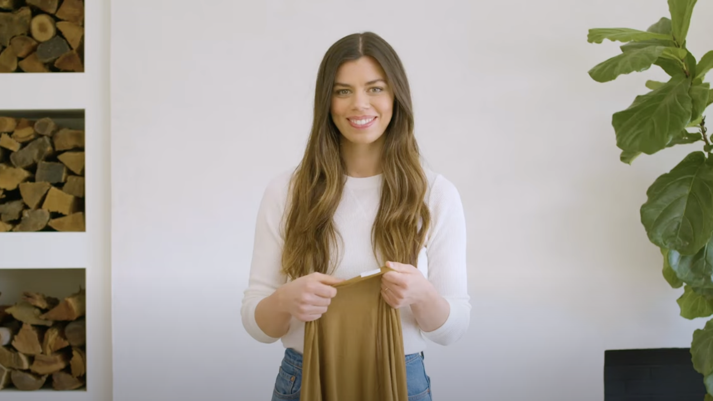 Woman wearing white sweater and blue jeans holding a folded olive green fabric in a bright room with wood and plant.