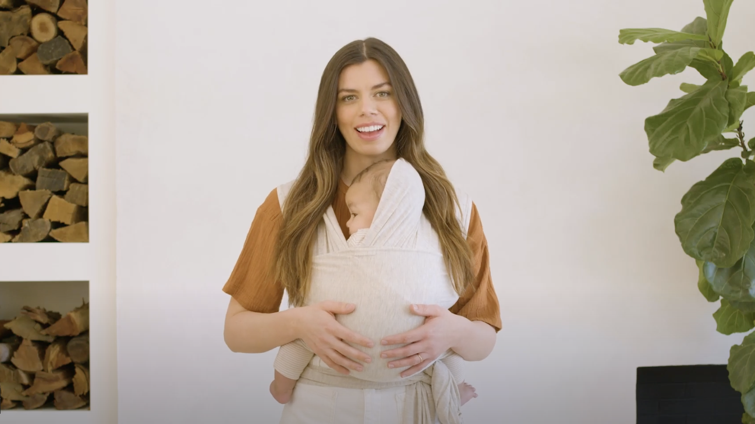 Woman wearing a beige baby wrap carrier holding a sleeping baby, standing indoors with stacked firewood and a green plant nearby.