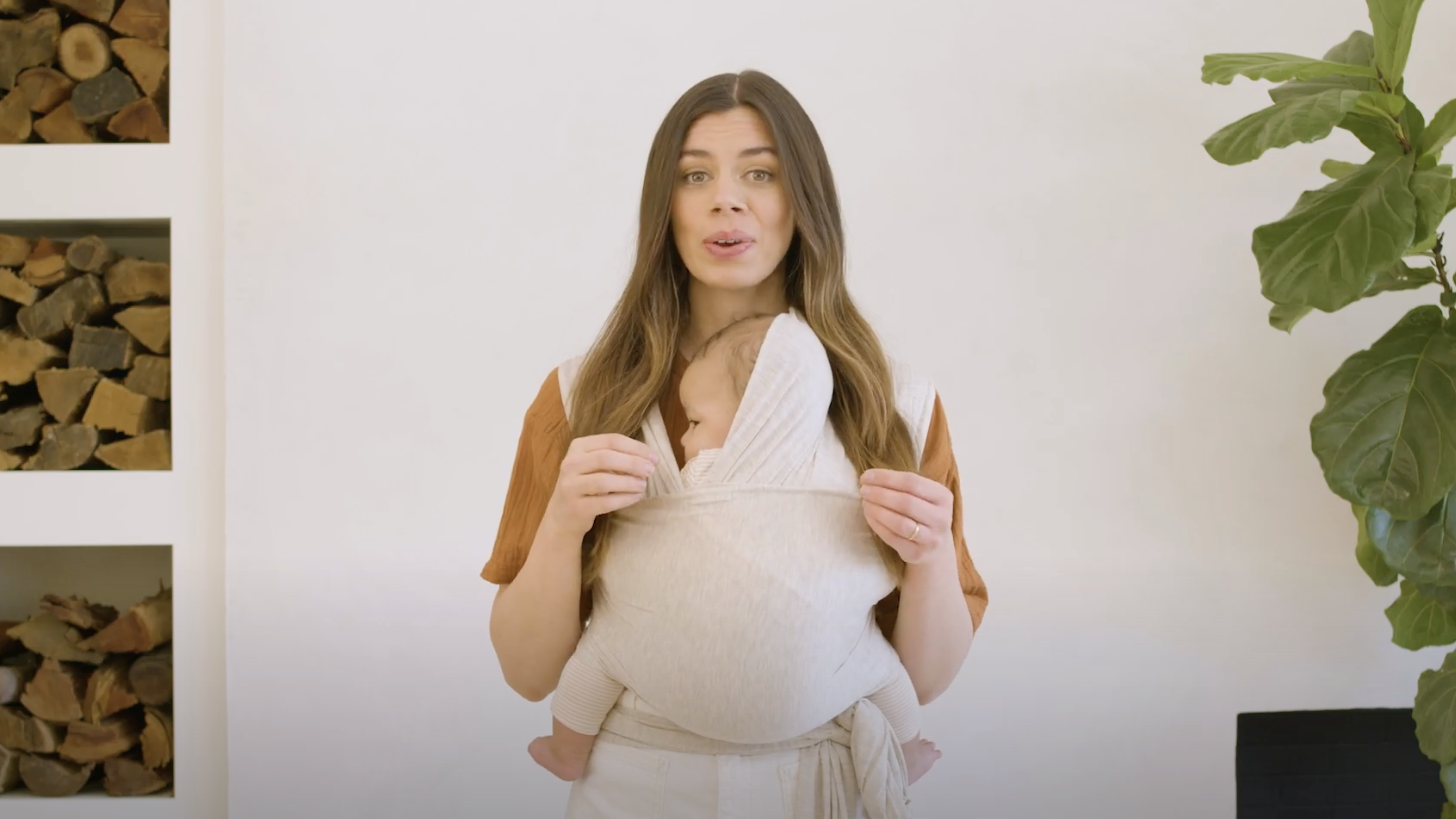 Woman wearing beige baby wrap carrier with hooded baby, standing indoors near stacked firewood and green plant.