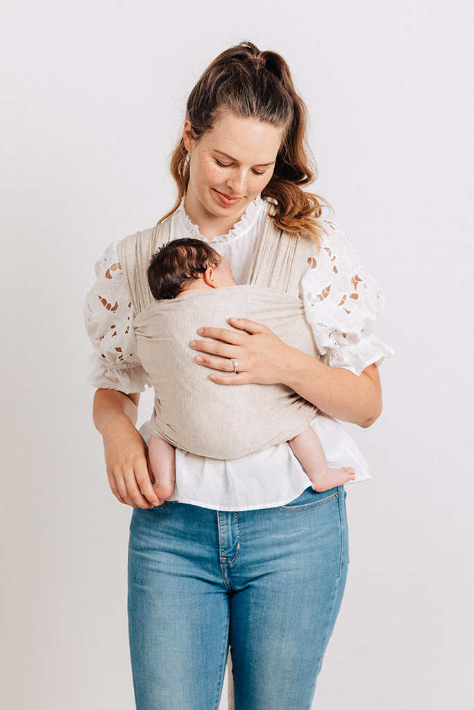 Beige soft fabric baby carrier wrap worn by woman holding infant facing inward, paired with white blouse and blue jeans.