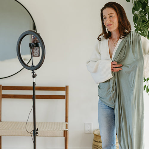 Woman holding sage green fabric or blanket, standing next to a ring light with a phone mounted on it.
