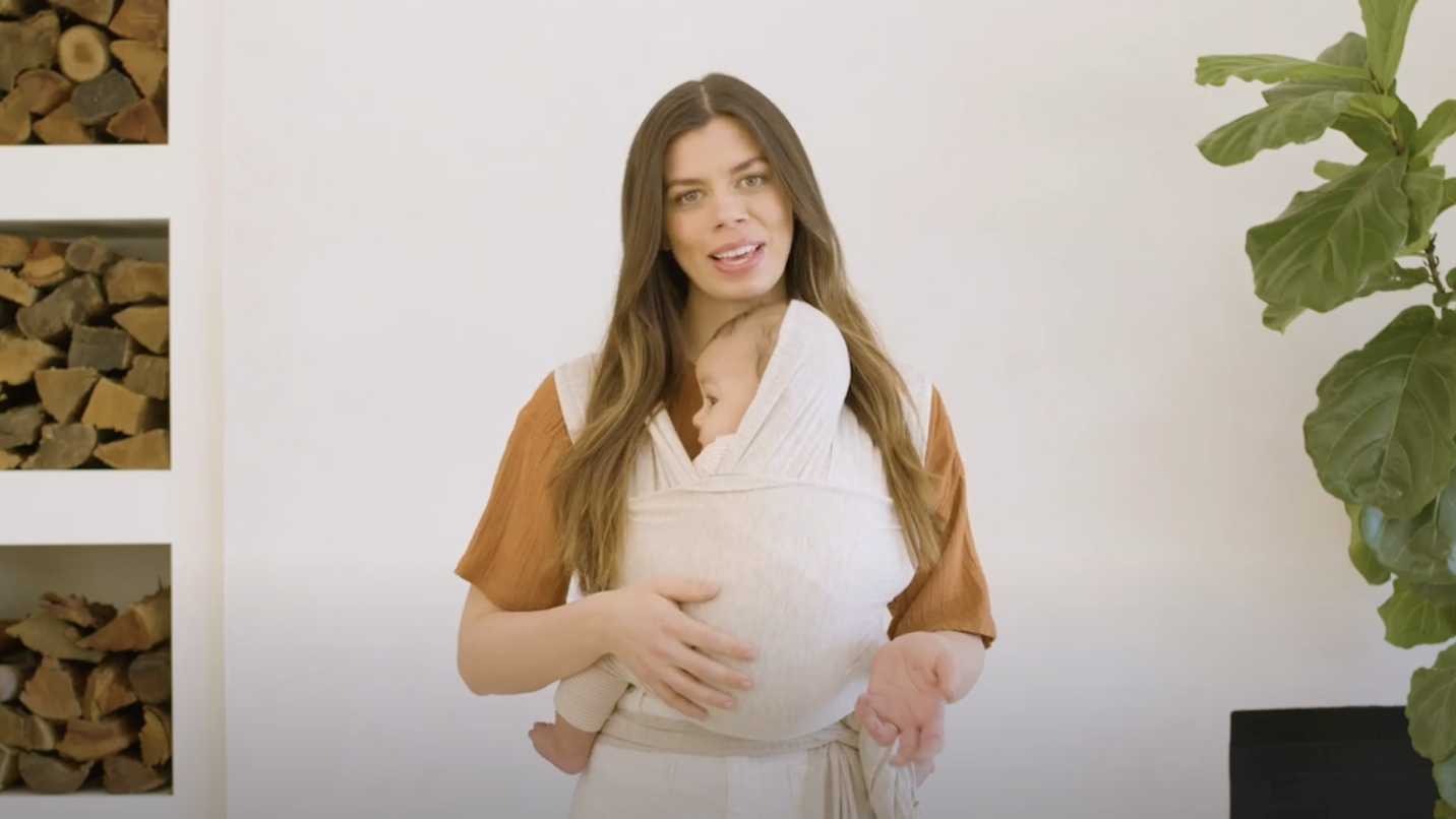 Woman wearing rust-colored top carrying baby in cream-colored hooded baby wrap carrier