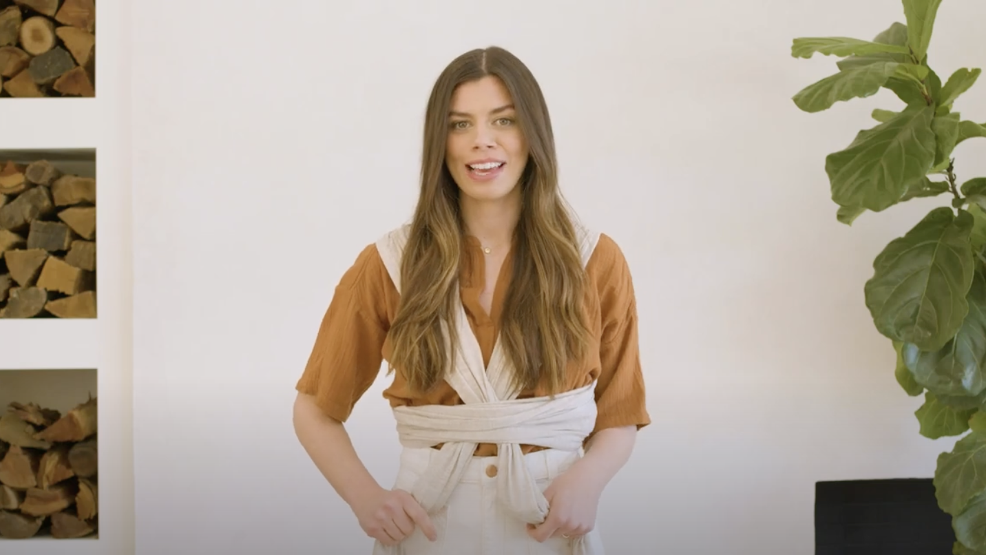 Woman wearing a rust-colored short sleeve shirt and beige wrap-around tied belt over high-waist pants, standing indoors.