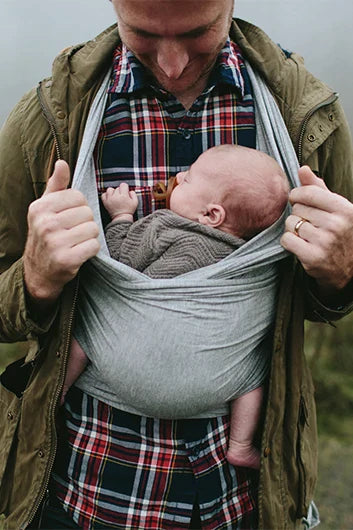 Man wearing green jacket and plaid shirt carrying baby in gray fabric sling carrier