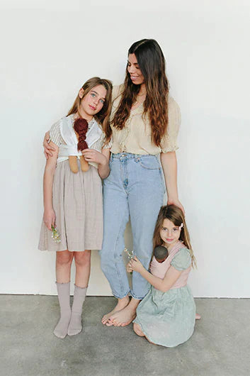 Woman in beige blouse and jeans with two girls, one in beige dress holding a doll, one in green dress holding flowers.
