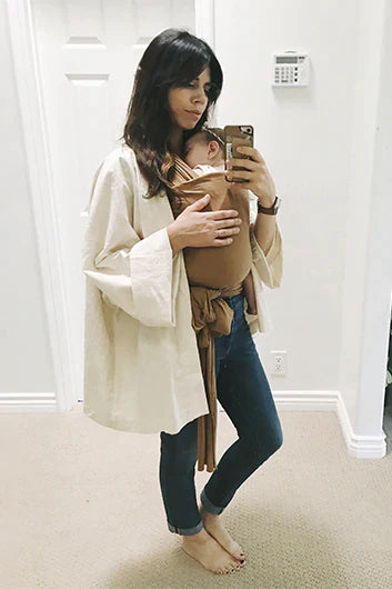 Woman wearing blue jeans and a beige loose shirt carrying a baby in a brown wrap carrier.
