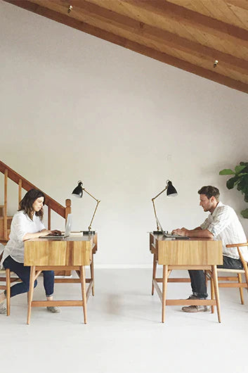 Two people sitting at adjacent wooden desks with black desk lamps, working in a minimalistic room.