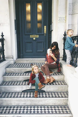 Woman in brown pants and plaid shirt holding baby in white hat, girl in red jacket and boots sitting on patterned steps, boy in blue shirt standing.