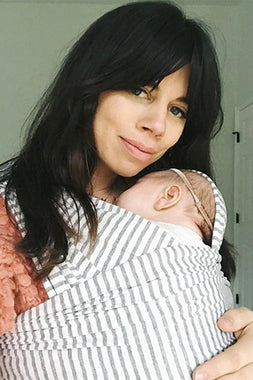 Woman wearing pink sweater carrying sleeping baby in white and gray striped fabric baby wrap.