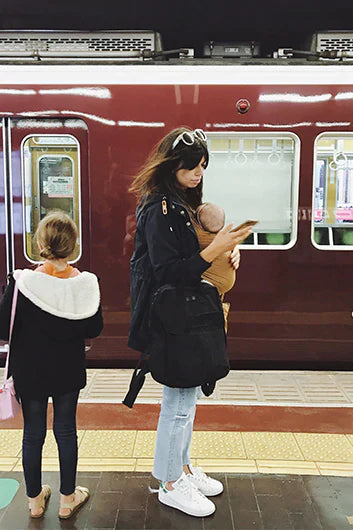 Woman in black jacket and light jeans holding baby in brown carrier, standing next to girl in black coat on train platform.