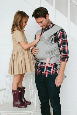 Gray baby wrap carrier worn by man holding baby, with girl in tan dress and purple boots standing on chair next to them.