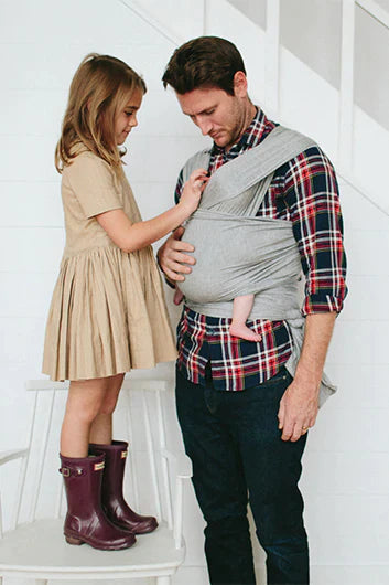Gray baby wrap carrier worn by man holding baby, with girl in tan dress and purple boots standing on chair next to them.